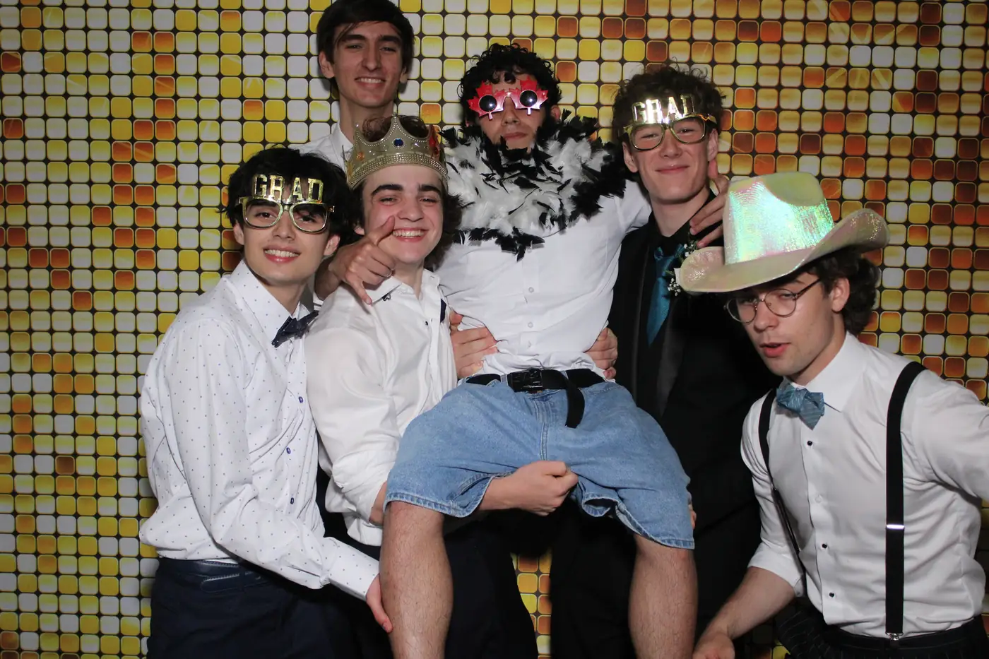 Un grupo de graduados con lentes de grad cargando a un amigo en el photobooth del prom
