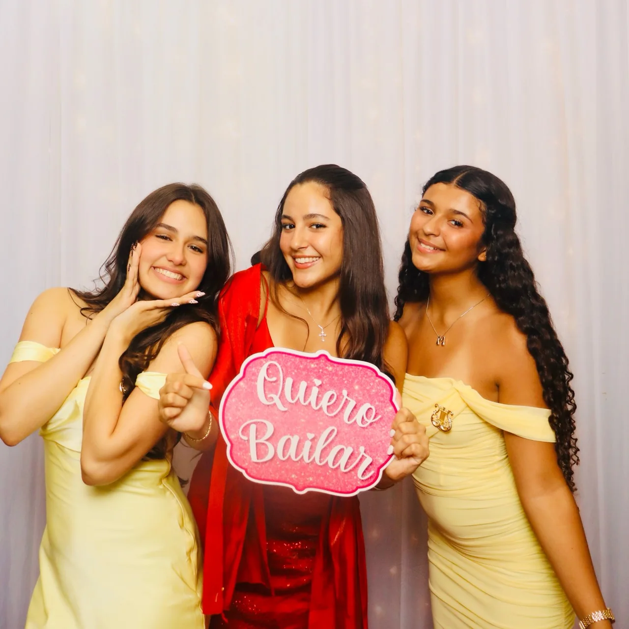 Three quinceañera guests in formal dresses holding a Spanish Quiero Bailar sign